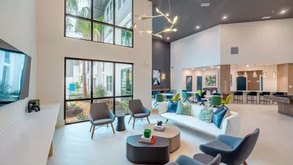 Image 1: Stylish resident lounge with modern furniture, floor-to-ceiling windows, and views of lush landscaping at Tropia Wellen Park Apartments.