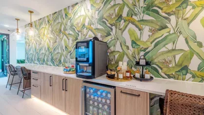 A vibrant coffee bar with tropical-themed wallpaper, illuminated by warm pendant lighting, offering a modern beverage station with snacks and refreshments.