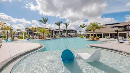 A spacious resort-style pool surrounded by lush landscaping, lounge chairs, and palm trees under a bright blue sky.