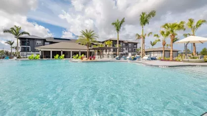 A spacious resort-style pool surrounded by lush landscaping, lounge chairs, and palm trees under a bright blue sky.
