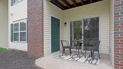 A cozy outdoor patio space with a small table, two chairs, and a patterned rug, overlooking a green yard.