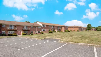 Wide parking lot and brick apartment buildings with balconies at Washington Gardens Apartments in Washington, NJ, under a bright blue sky.