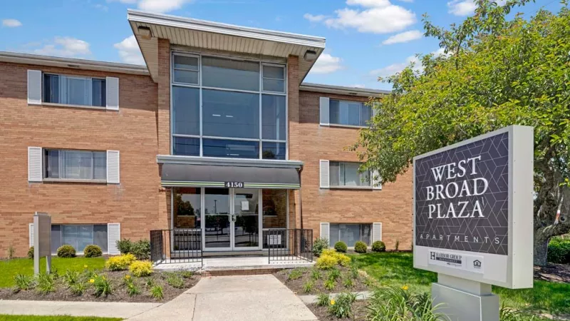 Brick apartment building with a glass entryway, surrounded by lush greenery and a 'Model Open' sign welcoming visitors.