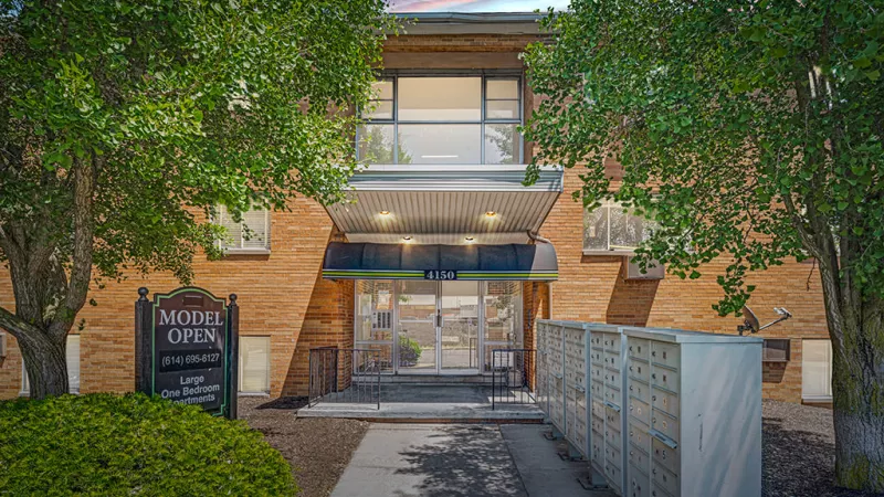 Brick apartment building with a glass entryway, surrounded by lush greenery and a 'Model Open' sign welcoming visitors.