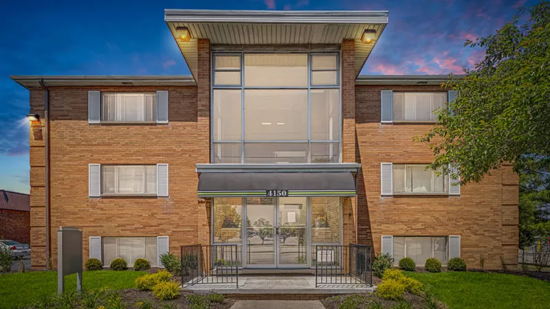 Brick apartment building with a glass entryway, surrounded by lush greenery and a 'Model Open' sign welcoming visitors.