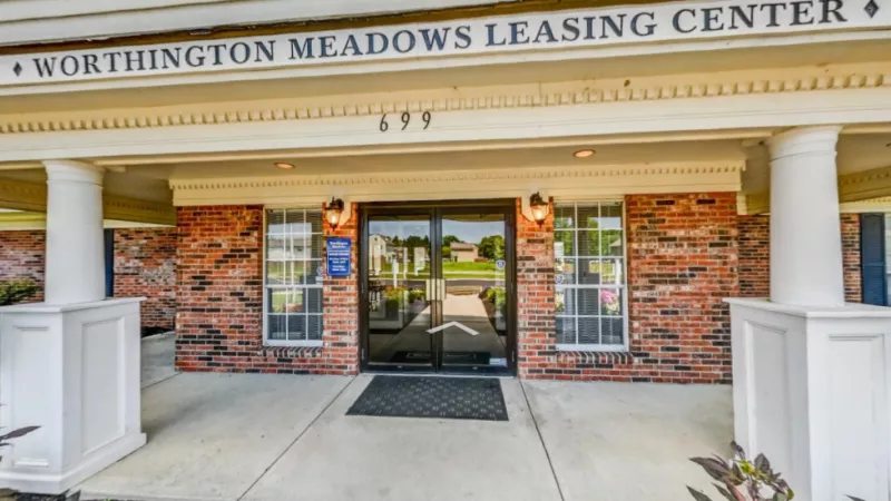 The Worthington Meadows Apartments leasing center features a welcoming brick exterior, large glass entry doors, and classic architectural columns. The entrance is surrounded by well-maintained landscaping, offering a warm and professional atmosphere for prospective residents.