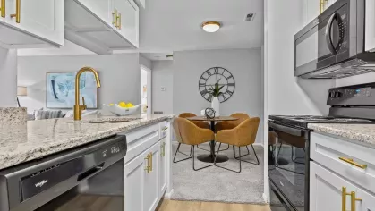 A contemporary kitchen with a gold faucet and white cabinetry, opening up to a dining area with brown chairs and a decorative wall clock.