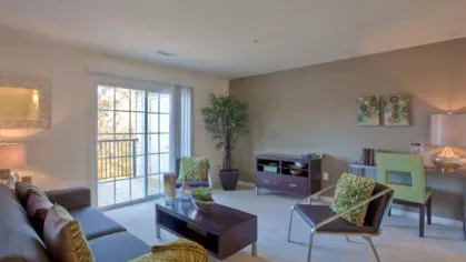 Open-concept living room with modern decor, featuring a cozy seating area, natural light from large windows, and a private balcony overlooking lush greenery.