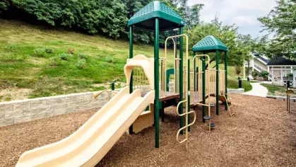 A children's play area at Vista At Town Green Luxury Apartments, featuring a playground with slides, climbing structures, and a mulch-covered surface set against a green hillside.