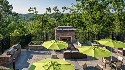 A stunning fireside lounge at Vista At Town Green Luxury Apartments, with outdoor seating, grilling stations, and green umbrellas offering a picturesque hillside view.
