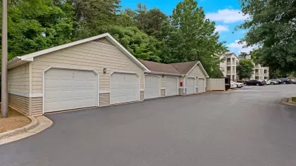 A row of detached garages with white doors, surrounded by lush greenery and apartment buildings in the background.