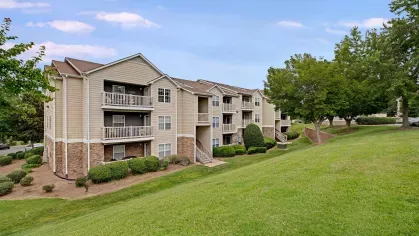 Three-story beige apartment building with balconies and well-maintained greenery on a gently sloping lawn.
