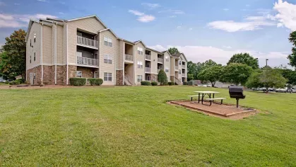 Outdoor picnic area with a grill and picnic table set against a beige apartment building and open green space.