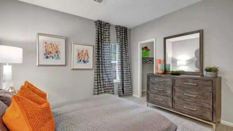 Bedroom with gray walls, orange accent pillows, colorful artwork, and a wooden dresser with a large mirror.