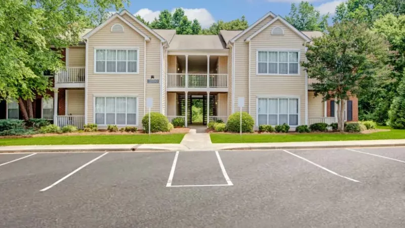Beige two-story apartment building with white trim and manicured landscaping in front of an empty parking lot.