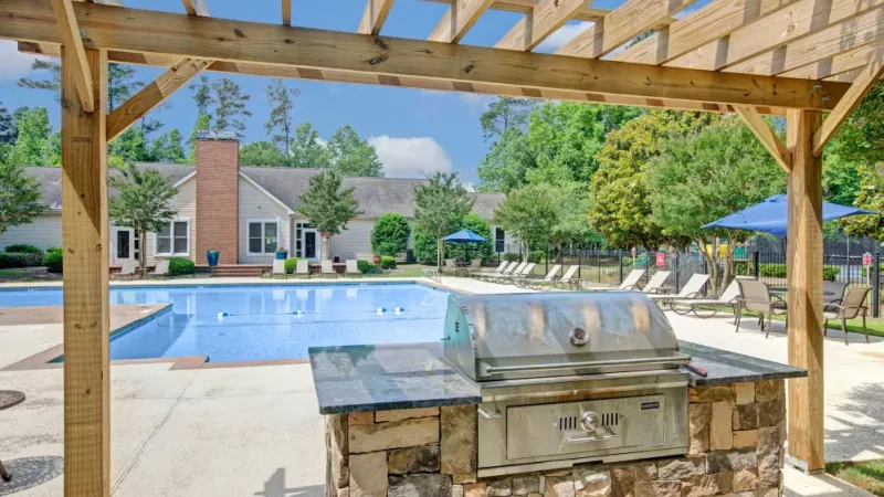 Outdoor grill station with a stone countertop under a wooden pergola next to a pool area with lounge chairs.
