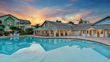 Resort-style pool with crystal-clear water reflecting a colorful sunset and surrounded by lush greenery and lounge chairs.