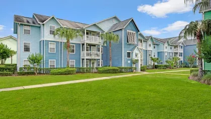 Blue multi-story apartment building with white balconies, surrounded by green lawns and palm trees.