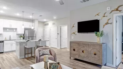 A modern open-concept living room with a ceiling fan, a mounted TV, and a kitchen featuring a sleek island with two white barstools.