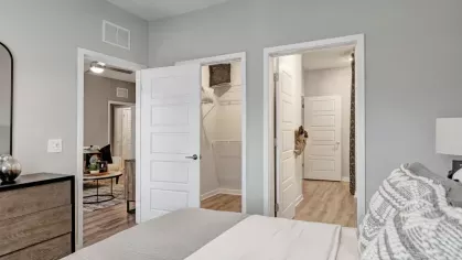 A bedroom with a neutral palette, featuring a bed with textured pillows, a ceiling fan, and open doors leading to a walk-in closet and bathroom.