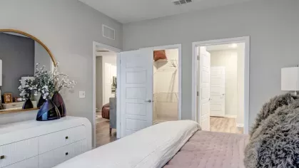 A bedroom with a soft color scheme, showcasing a neatly made bed, a white dresser, and an entryway to an en-suite bathroom.