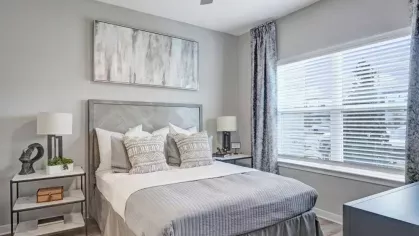 A chic bedroom with a gray upholstered headboard, coordinating bedding, and large windows framed by patterned curtains.