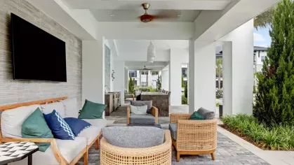 A covered outdoor lounge area with rattan chairs, plush cushions, a mounted TV, and a ceiling fan.
