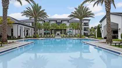 A luxurious swimming pool with crystal-clear water surrounded by palm trees and modern apartment buildings.