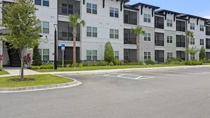 A modern three-story apartment building with white and gray exteriors stands next to a parking lot with a few parked cars.