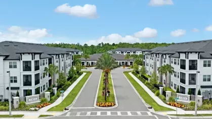 A bright daytime aerial view of the Aria community, with symmetrical palm-lined streets and modern apartment buildings.