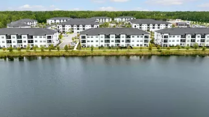 A panoramic lakefront view of the Aria apartment community, with white modern buildings reflecting on the water.