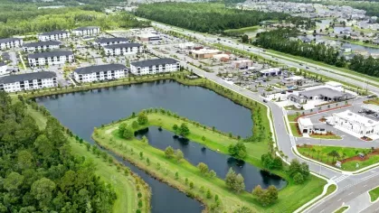 Aerial view of a modern apartment complex with multiple white buildings, a large pond, green spaces, and a nearby shopping center along a busy highway.