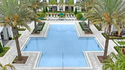 Expansive resort-style pool within a luxury apartment complex, framed by towering palm trees, lounge chairs, and shaded seating areas.