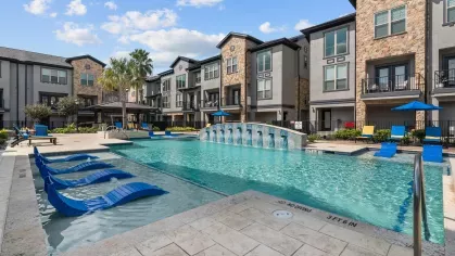 A stunning resort-style pool area with cascading water features, blue loungers, and shaded seating areas.