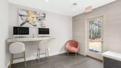 A cozy business center with two iMac computers, plush seating, and natural light streaming through the glass door at Aurella Cary Apartments.