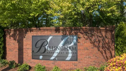 The Bellingham Apartment Homes sign is set against a classic red brick wall with vibrant greenery overhead.
