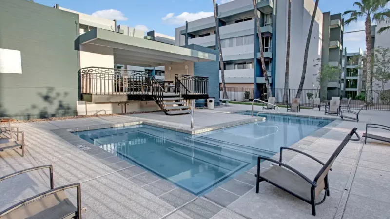 Outdoor pool area with a modern lounge space and a uniquely shaped pool surrounded by apartment buildings and palm trees.