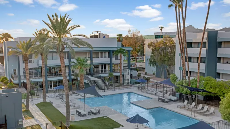 Aerial view of the apartment complex showing a large pool, lush greenery, and surrounding modern buildings.