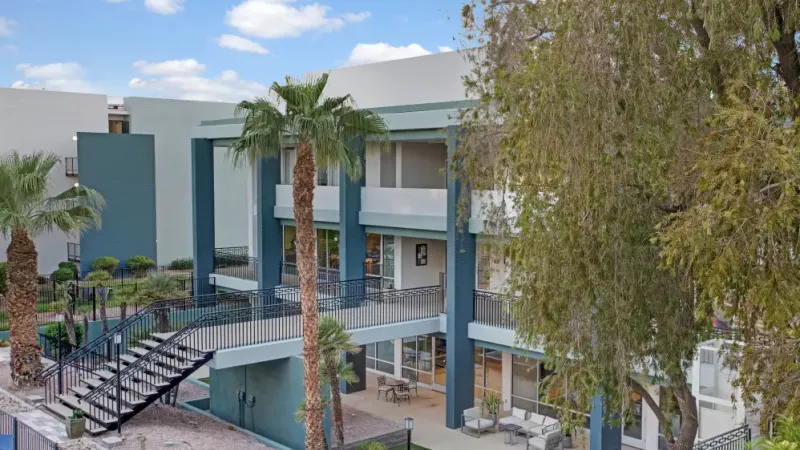 Exterior view of a modern apartment building with blue and white accents and lush palm trees.