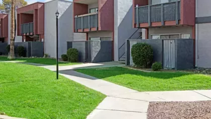 Sidewalks and green lawns in front of red and gray apartment buildings with private patios.