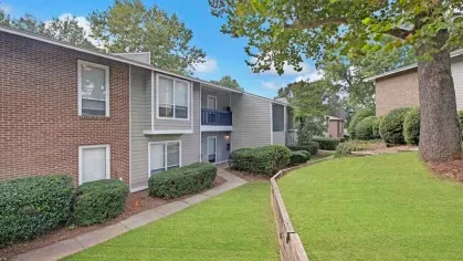 An apartment building with a mix of brick and siding, surrounded by lush greenery and mature trees.