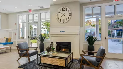 A welcoming seating area with a fireplace, large clock, and natural light from floor-to-ceiling windows.