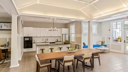 An inviting open-concept common area with a kitchen, long dining table, and cozy seating around a fireplace.