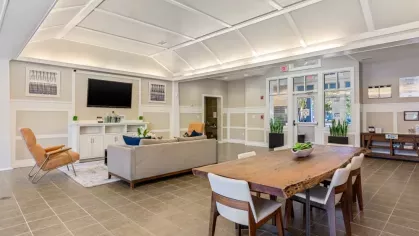 A modern communal lounge area with comfortable seating, a large wooden dining table, and a kitchen in the background.