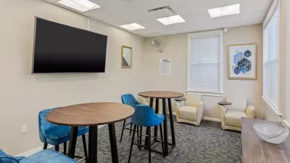 A cozy meeting room with a large wall-mounted TV, blue chairs, and small wooden tables.
