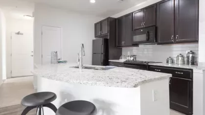 A kitchen with a curved granite island, two bar stools, and sleek cabinetry, leading to a front entry door.