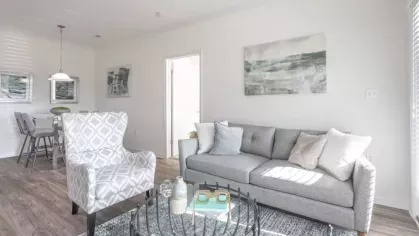 A cozy living room with a gray sofa, a patterned armchair, and large windows letting in sunlight.
