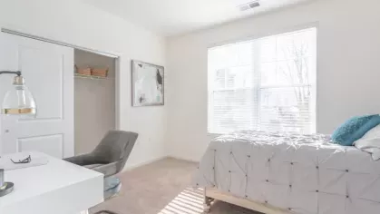 A bright bedroom featuring a cozy bed with a gray tufted comforter, a desk with a modern chair, and large windows allowing natural light to illuminate the space.