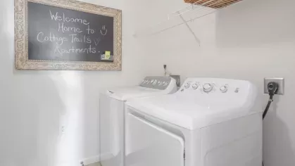 A laundry room equipped with a washer and dryer, an open shelf above, and a decorative chalkboard sign that reads "Welcome Home to Cottage Trails Apartments."