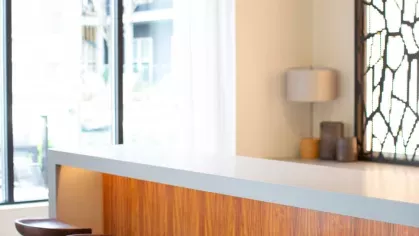 A modern kitchen island with a sleek white countertop, wooden base, and two brown leather stools, accented by a decorative lamp and large windows.